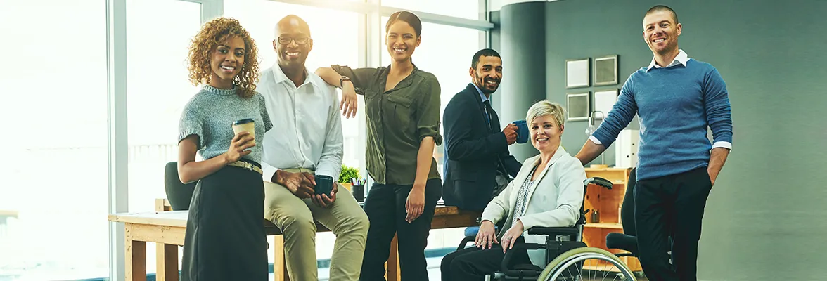 Group of six diverse professionals smiling in an office environment, one of whom is using a wheelchair.